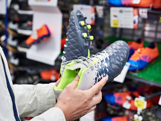 A person holding a pair of grey and lime green football boots, examining the design and studs in a sports store.