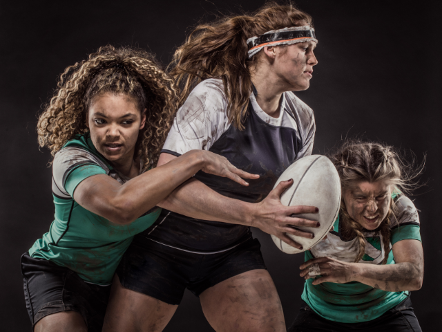 Three female rugby players battling for the ball in a tense match.