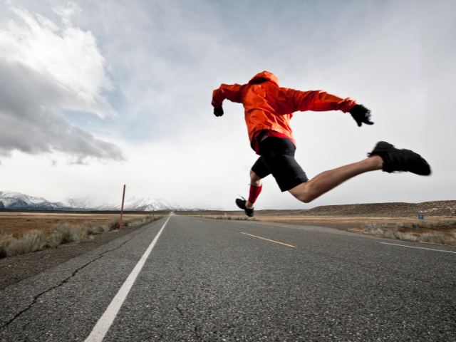 A runner in a red jacket leaping mid-air on an empty road with mountains in the background.