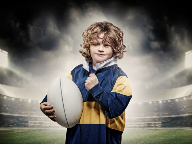 A young rugby player holding a rugby ball, standing in a stadium.