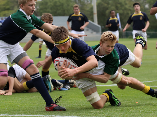 Rugby player in a dark jersey dives to score a try while being tackled by a player in green, with teammates rushing in to support.