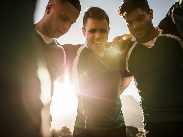 Rugby players standing close together in a huddle, with sunlight shining between them, reflecting their camaraderie.