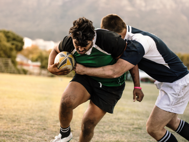 Rugby player carrying the ball while being tackled from behind during an outdoor match.