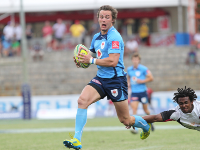 A rugby player in light blue running with the ball during a game, while an opponent in dark-colored jersey attempts to tackle him from behind.