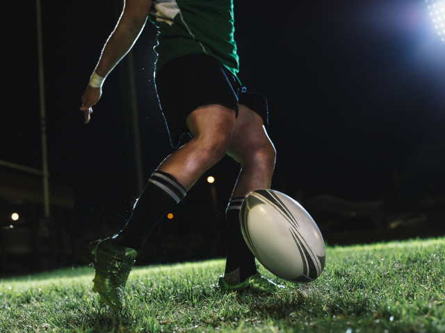 A rugby player in green and black jersey kicks the ball during a night game on a well-lit field. 