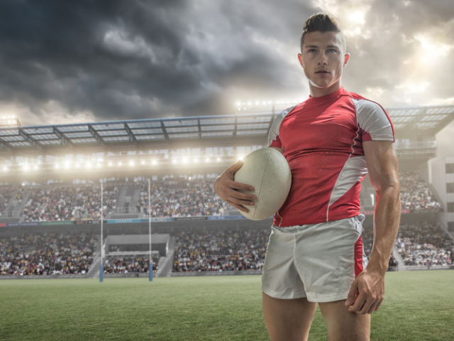 A rugby player in a red and white uniform holding a rugby ball, standing confidently on the field with a stadium full of spectators in the background.