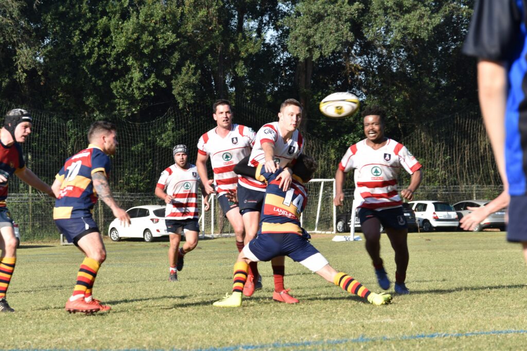 Rugby players in action during a match. One player in a striped red and white jersey is being tackled by an opponent in a multicolored jersey, while other players surround them, preparing for the next move.