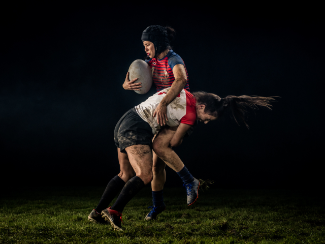 Female rugby player being tackled while carrying the ball during a nighttime match.