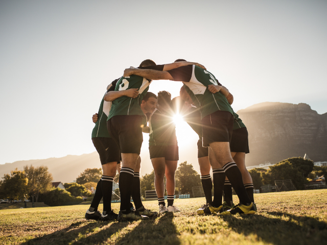 A team of rugby players huddled together on the field with the sun setting in the background, creating a powerful silhouette.