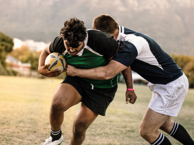 Rugby player carrying the ball while being tackled by another player on the field.