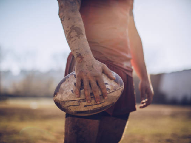 A rugby player stands holding a ball with mud-covered hands.