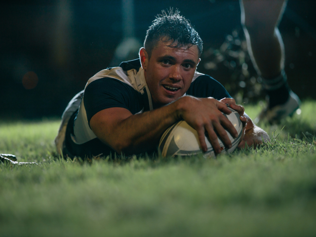 A rugby player smiling while grounding the ball for a try during a night match.