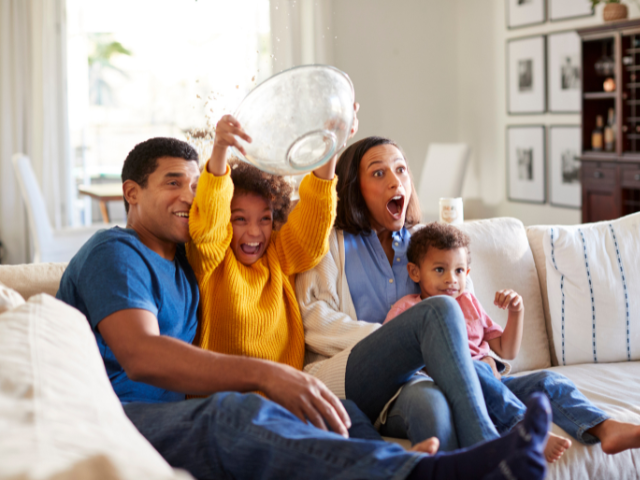 A family sitting on a couch, excitedly watching something on TV, with one of the children holding a bowl above their head.