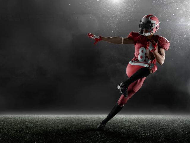 A rugby player in a red uniform making a powerful kick on a dark stadium field.