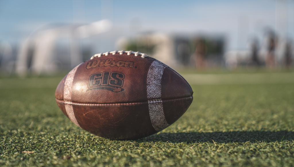 A close-up of a Wilson CIS rugby ball resting on a grassy field under clear skies.