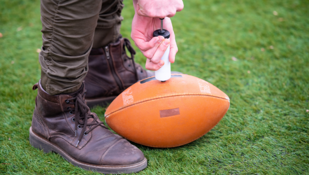 A person inflating a rugby ball using a hand pump on a grassy field.