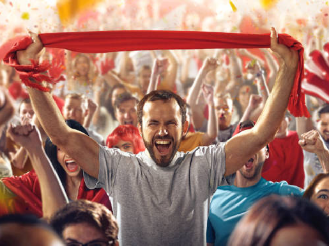 A man cheering in a crowd, holding a red scarf at a sports event.