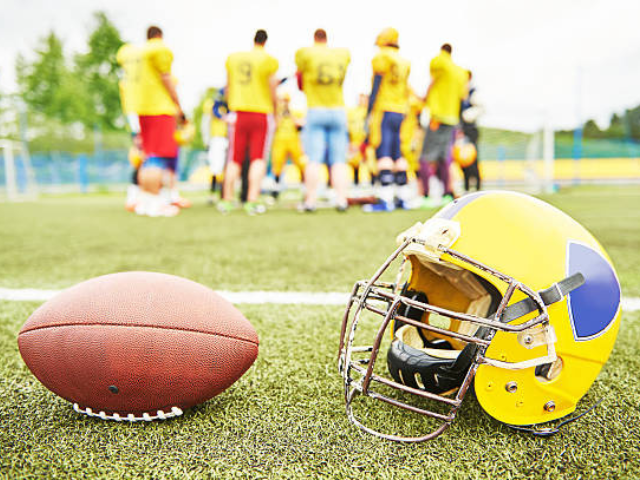 A football helmet and ball placed on the grass with players standing in the background.