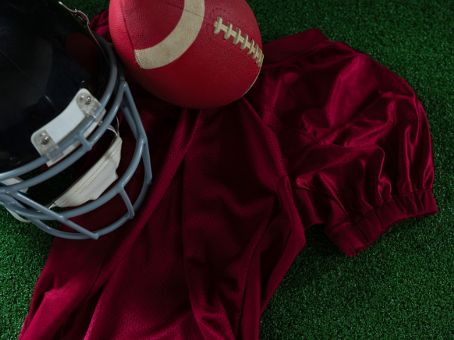 A football helmet, ball, and red jersey laying on a green turf field.