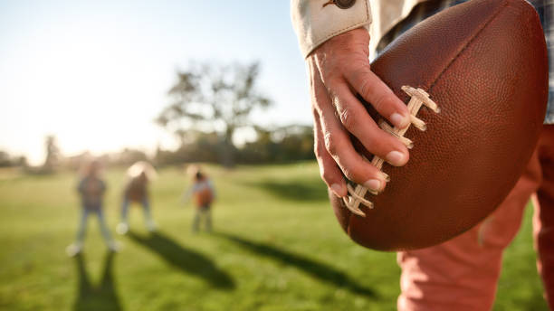 Close-up of a hand holding a rugby ball with kids playing in the background.