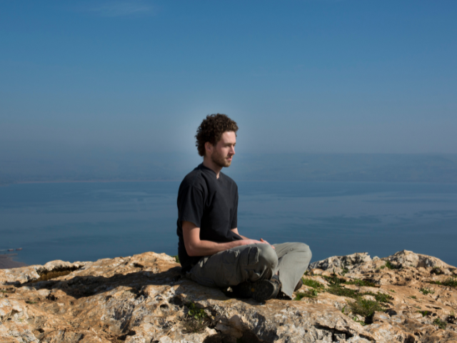 A man sitting on a rocky hilltop meditating with a scenic view in the background.