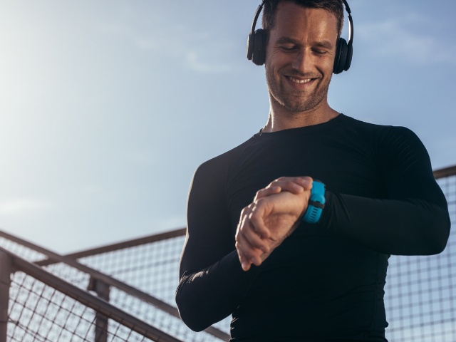 A man checking his sports watch while smiling, wearing headphones and a long-sleeved athletic top.