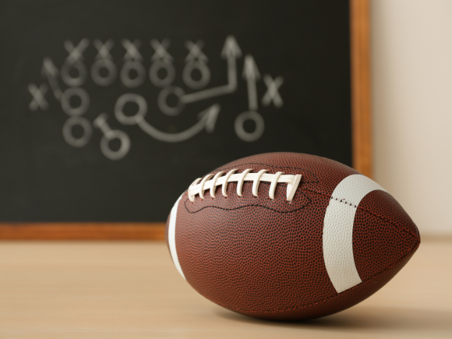 A traditional brown football sitting in front of a chalkboard with a game strategy drawn on it.