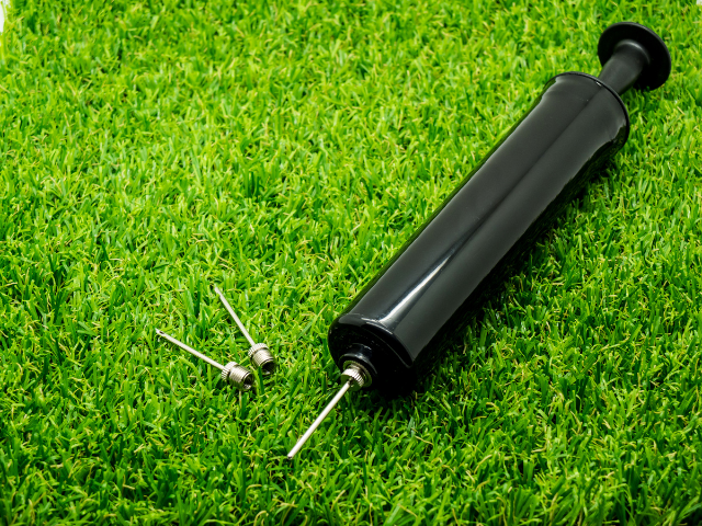 A black ball pump with two inflating needles placed on green artificial grass.