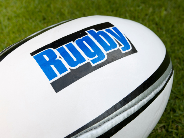 A white rugby ball with "Rugby" written on it in bold blue letters, placed on grass.