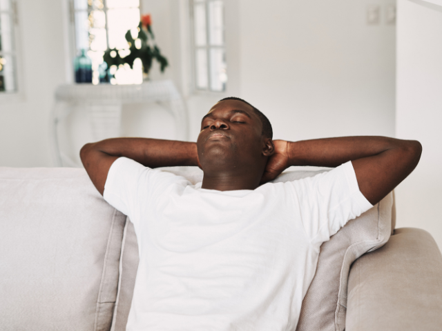 A man resting on a couch with hands behind his head, eyes closed, and appearing relaxed.