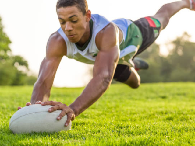 A rugby player diving to score a try, with arms outstretched and the ball inches from the ground.