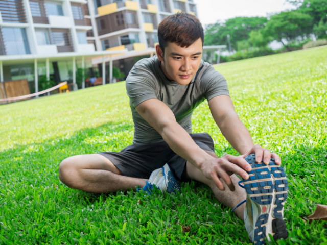 A man sitting on the grass, stretching his leg with a gentle pull towards his body.