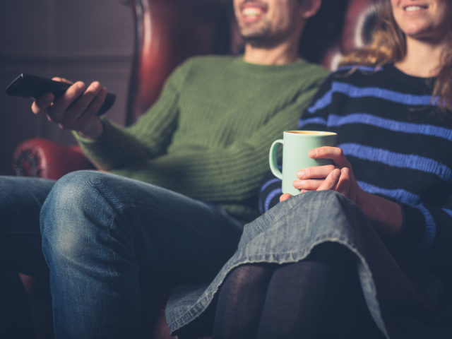 A couple sitting on a couch watching TV while holding a mug.