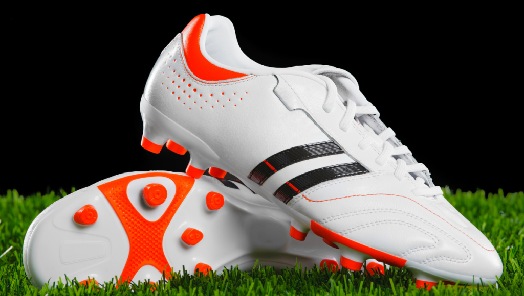 A sleek white rugby boot with black and orange accents placed on green turf.