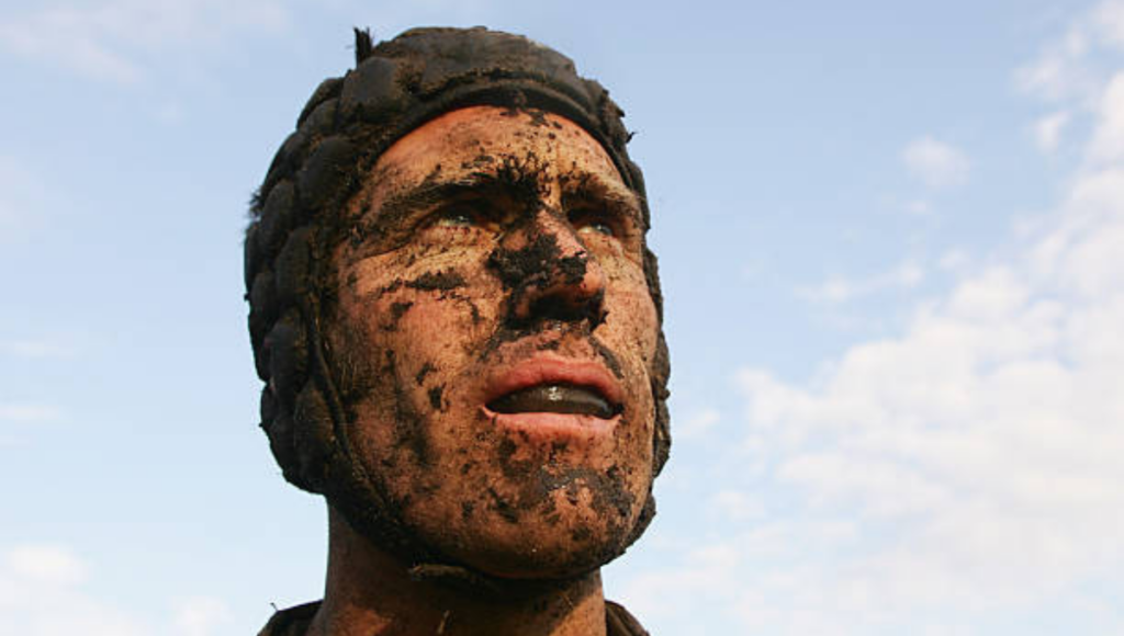 A rugby player’s face covered in mud, looking focused and determined.