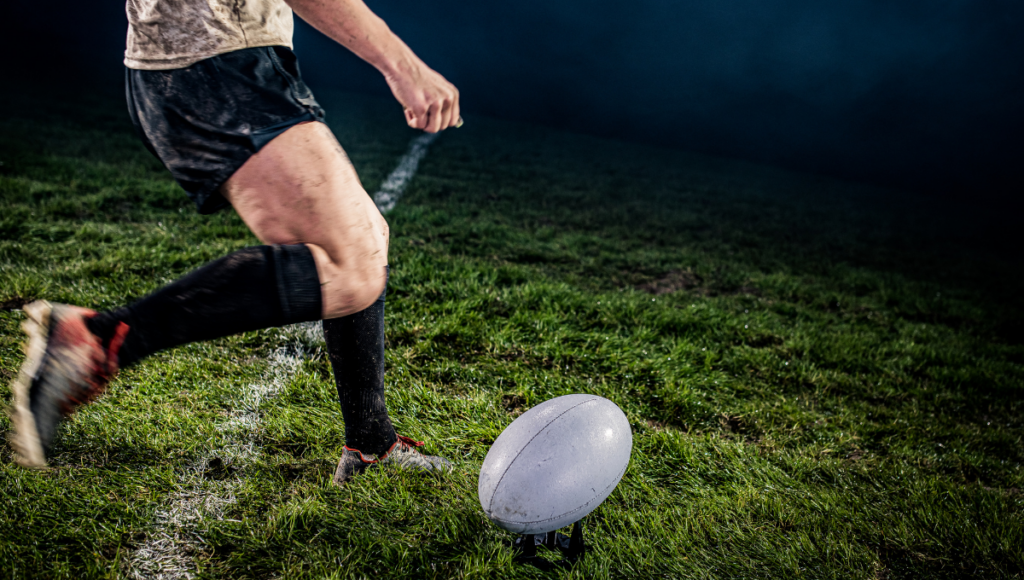 Rugby player kicking a ball from a tee under stadium lights.