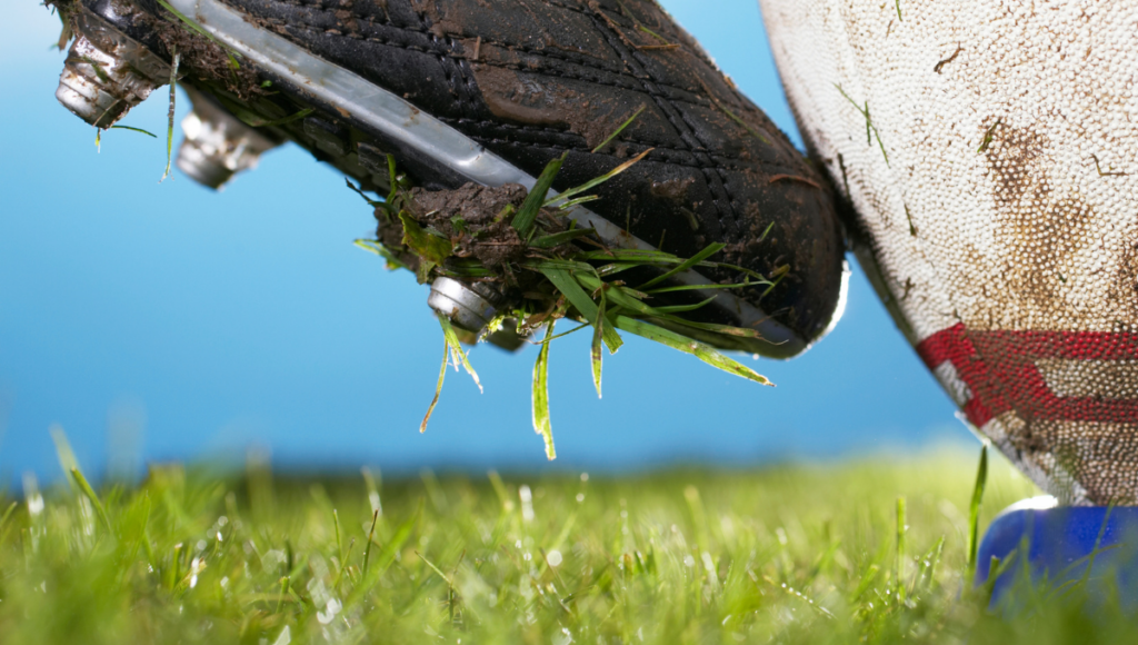 A close-up image of a muddy rugby boot about to kick a ball on a grassy field.