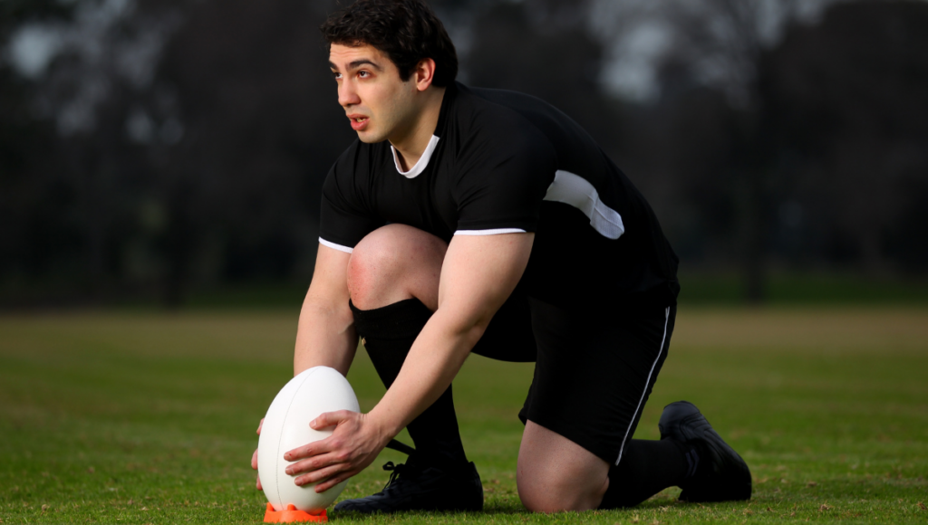 A rugby player kneeling on a field, preparing to kick a rugby ball placed on an orange tee.