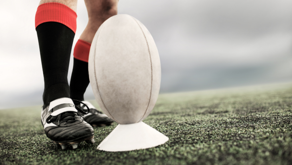 Close-up of a rugby ball on a tee as a player prepares to kick it, focusing on the ball and tee.