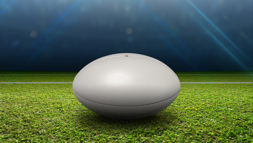 A rugby ball resting on a grassy field under stadium lights with a dark sky in the background.