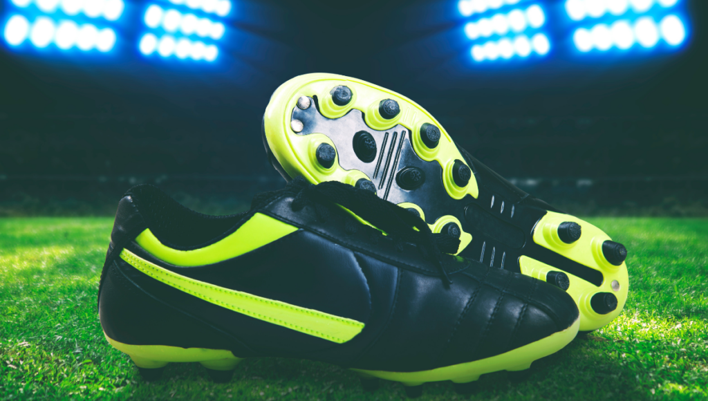 A pair of black and neon yellow rugby boots with studs on the sole, lying on the grass under stadium lights.