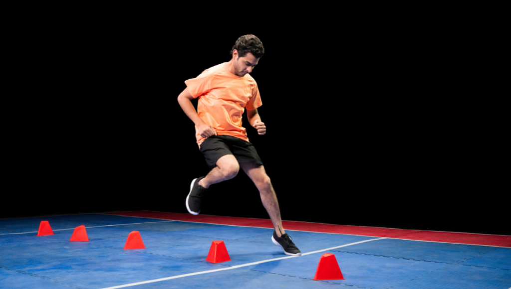 A man in an orange shirt performing agility drills with red cones arranged in a line on a blue indoor court.