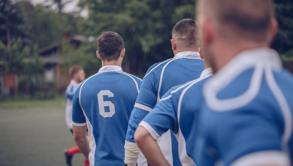 A team of rugby players in blue jerseys, lined up with their backs to the camera, ready for a match in a light drizzle.