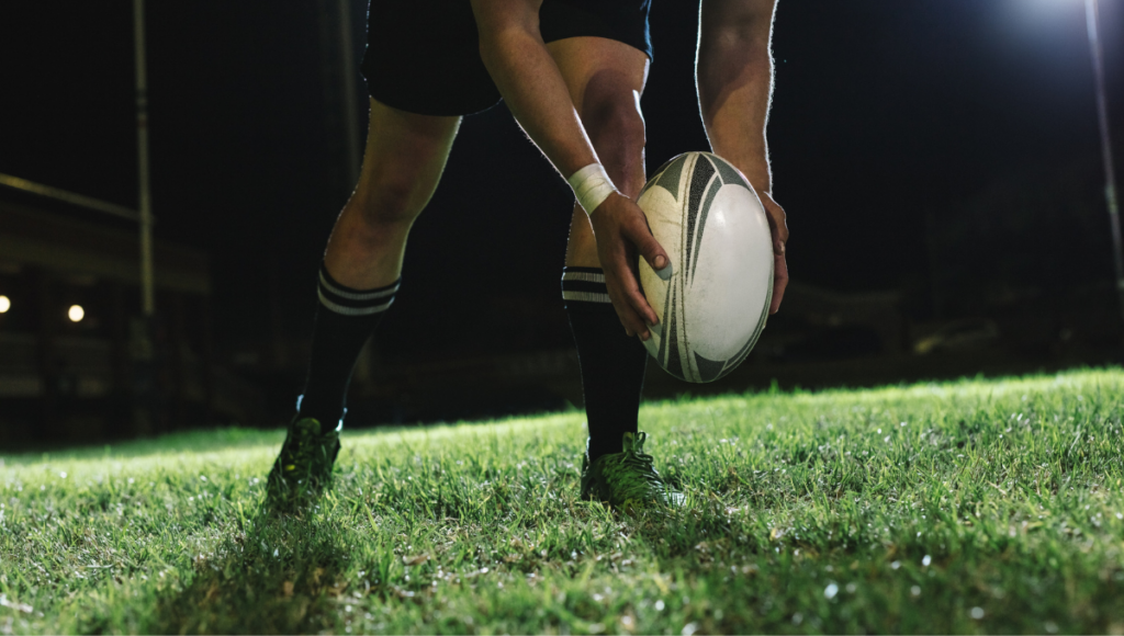 A rugby player crouching under the bright stadium lights, preparing to pass a rugby ball during a nighttime practice or match.