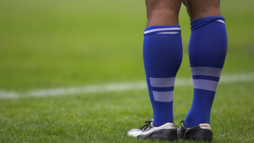 A person standing on a grassy field, wearing blue socks with white stripes and black sports shoes.