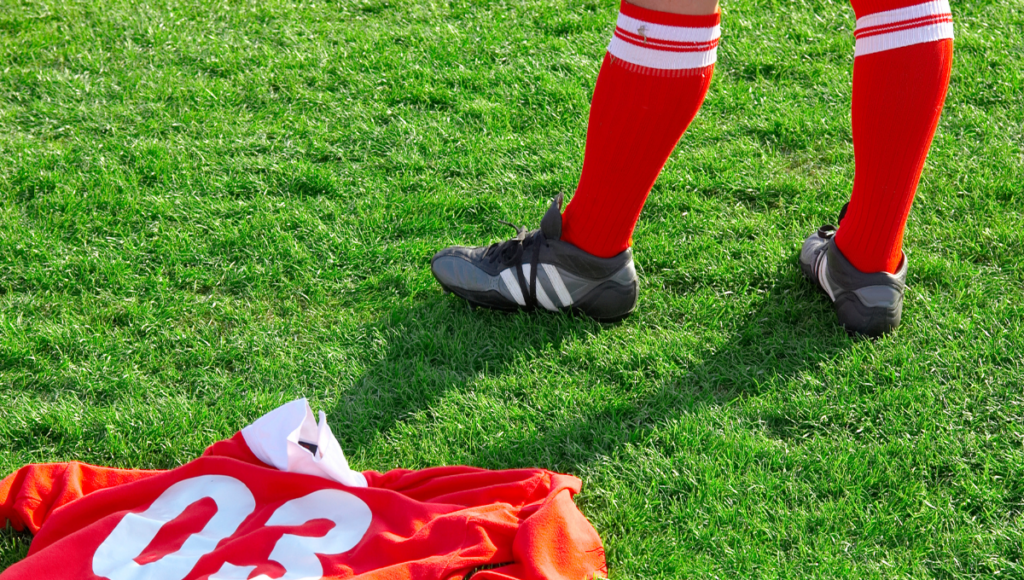 A soccer player standing on a grassy field wearing red knee-high socks with soccer cleats, with a red soccer jersey number 03 on the ground.