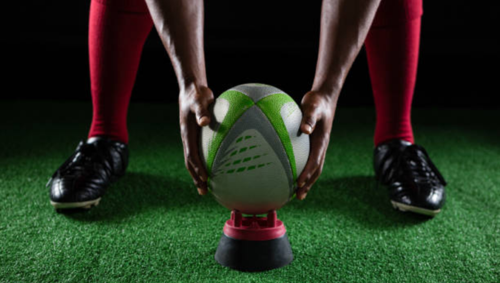 A rugby player placing a green and white rugby ball on a kicking tee, wearing black cleats and red socks, with a green artificial turf field in the background.