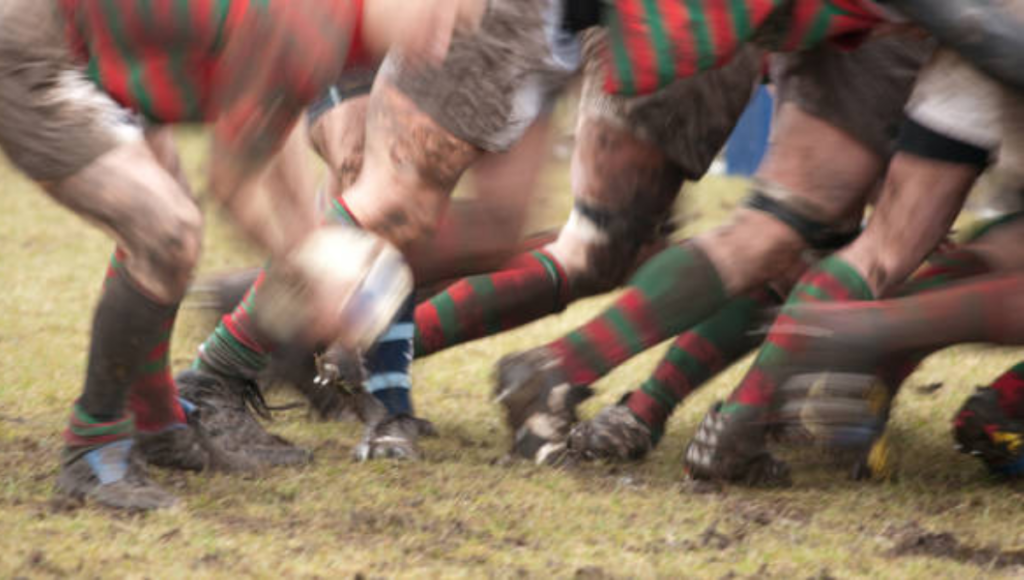 A blurred action shot of a rugby scrum with players in green and red socks on a muddy field, competing for the ball.