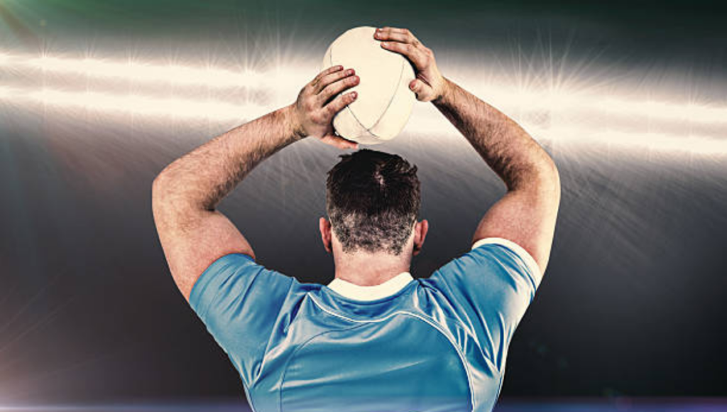 A rugby player seen from the back, in a blue jersey, holding a rugby ball above his head as if preparing to throw it during a game under stadium lights.