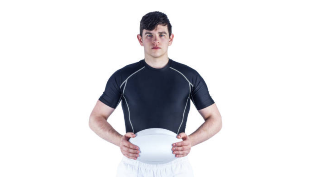 A rugby player in a black sports shirt and white shorts, holding a rugby ball, standing in front of a white background.